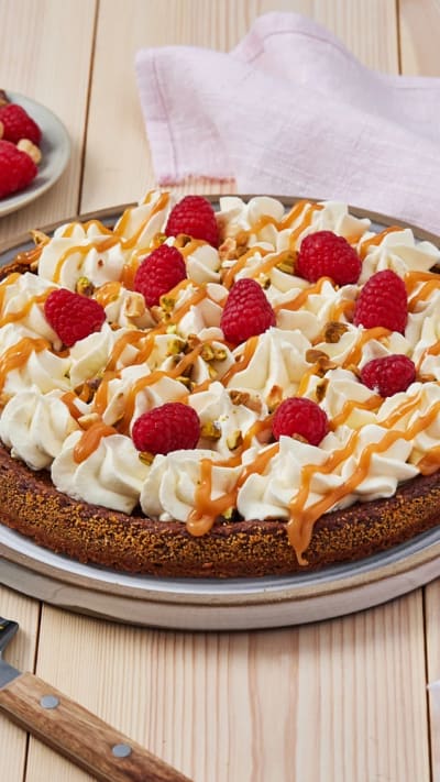 En kladdkakecheesecake står på ett bord med vispad grädde, bär och nötter.