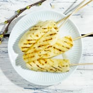 En tallrik med grillad ananas på pinnar står på en träyta.