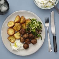 En tallrik med köttbullar, stekta potatisar, sallad och en klick gräddfil serveras bredvid bestick och ett glas med isbitar.