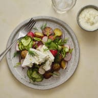 En tallrik med sallad bestående av grönsaker och potatis, serverad med en vit sås och bestick på ett bord.