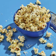 En skål med popcorn står på en blå bakgrund bredvid ett rivjärn och utspridda popcorn.