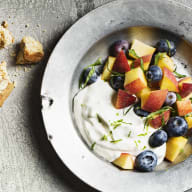 En bakplåt med ett potatisgratin och en tallrik med yoghurt och frukt står på en grå yta bredvid några brödskivor.