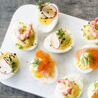 En vit serveringsbricka med små portioner av olika förrätter och en gaffel ligger på en grå yta.