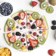 En fruktpizza med jordgubbar, kiwi och andra frukter ligger på ett bord tillsammans med skålar av bär och fruktbitar.