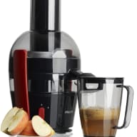 En elektrisk juicepress står bredvid en behållare med färskpressad juice och några halverade äpplen på en reflekterande yta.