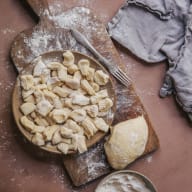 En träskärbräda med hemgjorda pasta, en gaffel, en degklump och en skål med mjöl ligger på en yta bredvid en kökshandduk.