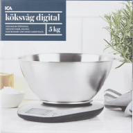 En digital köksvåg med en stor metallskål står på en yta bredvid en kruka med örter.