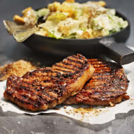 Två grillande köttbitar ligger på ett papper bredvid en skål med sallad på en mörk yta.