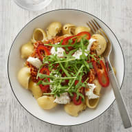 En tallrik med pasta, grönsaker och sås står på en ljus yta bredvid en gaffel.