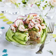 En tallrik med sallad bestående av gurka, räkor och rädisa står på ett dukat bord med blommor i bakgrunden.