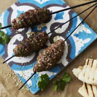 Grillspett med kött ligger på en tallrik bredvid pitabröd och en skål med kryddor.