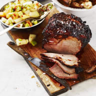 En skiva grillat kött ligger på en skärbräda bredvid en skål med potatissallad och en tallrik med bröd.