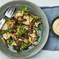 En tallrik med sallad bestående av svamp, grönsaker och nötter står bredvid en skål med sås på ett bord.
