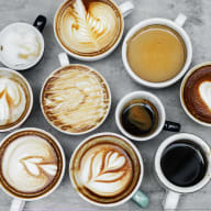 Flera koppar med olika kaffedrycker står på en grå yta, några med mönster i skummet.