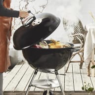 En person grillar mat på en rund grill utomhus bredvid ett dukat bord med växter och tillbehör.