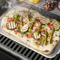 En pizza med grönsaker tillagas på en grill med örter, citroner och kronärtskockor bredvid.