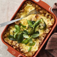 En ugnsform med kycklinglasagne med broccoli, garnerad med färska blad, och en sked ligger i maträtten.