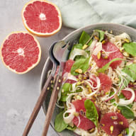 En tallrik med sallad innehållande grapefruktbitar, lökringar och gröna blad serveras bredvid halvor av grapefrukt och bestick på en duk.