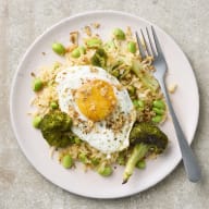 Ett stekt ägg ligger på en tallrik med stekt ris, broccoli och bönor. Rätten är toppad med rostad lök och på tallriken ligger en gaffel.