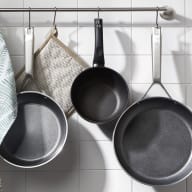 En köksvägg med upphängda stekpannor, en handduk och en grytlapp på en metallstång.