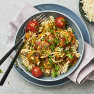 En tallrik med pasta, tomater och grönsaker står på ett bord bredvid bestick och en servett.