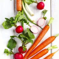 Färska grönsaker ligger utspridda på en vit träyta, inklusive morötter, rädisor och olika bladgrönsaker.
