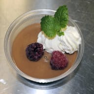 En dessert i en genomskinlig skål innehåller pudding, grädde, bär och ett mintblad.