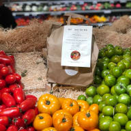 En papperspåse med etikett står bland högar av tomater och paprikor på hö i en butik.