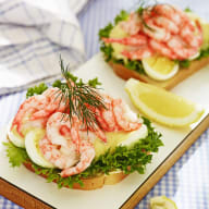 Två smörgåsar med räkor, sallad och citron ligger på träskärbrädor bredvid en rutig duk.