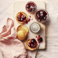 Körsbärssylt i burkar. Bredvid scones med sylt på. Rosa servett på sidan.