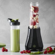 En blandare med frukt och grönsaker står bredvid en smoothieflaska och några hela frukter på en bänkskiva.