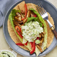 En tallrik med en öppen wrap innehållande kött, grönsaker och sås står på ett bord bredvid en skål med extra sås och en gaffel.