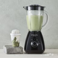 En mixer med grön vätska står på en bänk bredvid en liten behållare med något vitt i.