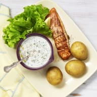 Stekt lax, kokt färskpotatis, ett salladsblad och en skål med östsås som ligger på en tallrik. Tallriken står på en gul servett.
