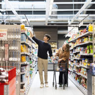 Två personer i en mataffärsgång där en sträcker sig efter en vara medan den andra håller i en kundvagn.