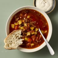 En skål med soppa, en sked, en brödbit och en liten skål med sås står på ett underlag.