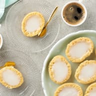 En tallrik med små bakverk och två koppar kaffe står på ett bord tillsammans med en servett och en sked.