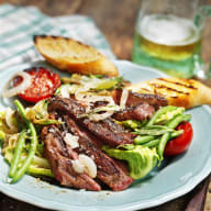 En tallrik med grillat kött, sallad och bröd står på ett träbord bredvid ett glas dryck.