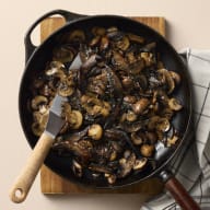 En stekpanna med svamp står på en skärbräda bredvid en kökshandduk.