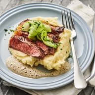 En tallrik med köttstycke och potatismos står på ett bord bredvid ett vattenglas och en skål med sallad.