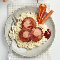 En tallrik med skivad korv på pasta, bredvid en gaffel och några grönsaker.