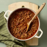 En gryta med köttfärssås står på en skärbräda bredvid en kökshandduk.