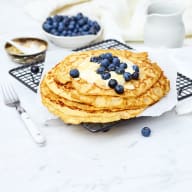 En hög med pannkakor med blåbär på toppen ligger på ett galler bredvid en skål med fler blåbär och en gaffel.