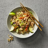 Pad thai med räkor i en skål som toppas med salladslök, jordnötter och limeklyftor. I skålen finns ätpinnar.