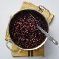 En kastrull med svart ris med en sked i som står på en duk.