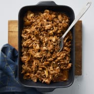 En ugnsform med pulled pork som står på en skärbräda bredvid en duk, i ugnsformen finns en sked.