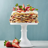 En tårta med flera lager står på ett vitt kakfat och är dekorerad med jordgubbar och gröna blad på toppen.