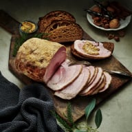 En skärbräda med skivor av kött och bröd, omgiven av tallrikar med nötter och örter.