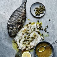 En grillad fisk ligger på en yta bredvid en citron, en tallrik med kapris och en skål med sås.
