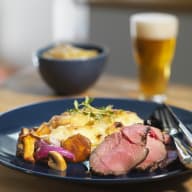 En tallrik med kött, grönsaker och gratäng står bredvid ett glas öl och en skål på ett bord.